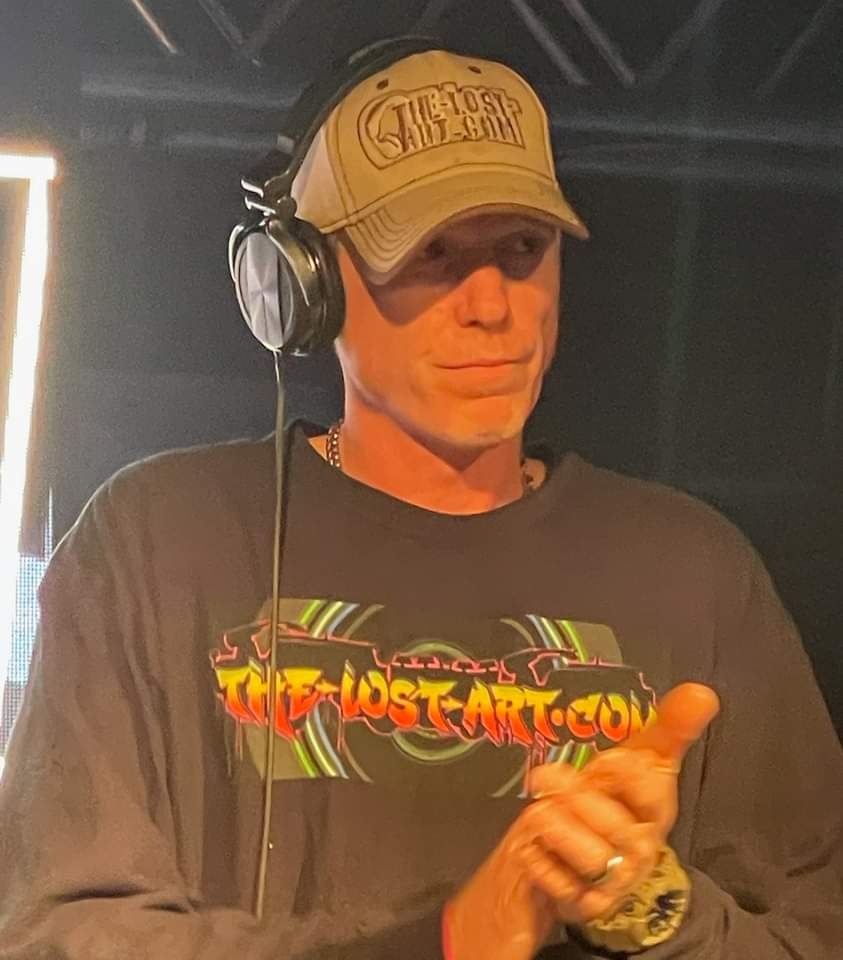 DJ Natural Nate® in The-Lost-Art.com long sleeve and TLA hat with headphones, thumbs up holding a coin under warm stage light